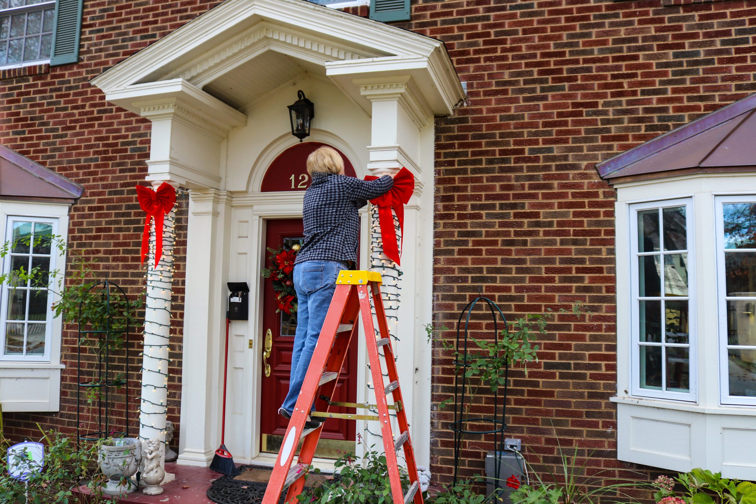 woman using ladder to hang holiday decorations on outside of house